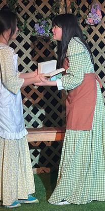 Two students in long gingham dresses hold a book between them on stage